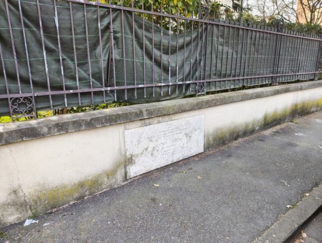 Lapide a Carta in un contesto urbano, parzialmente coperta da una rete e circondata da vegetazione.