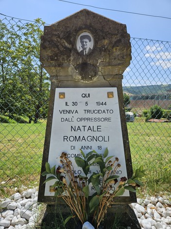 Cippo commemorativo dedicato a Natale Romagnoli, con foto e decorazioni floreali, situato in un'area verde.