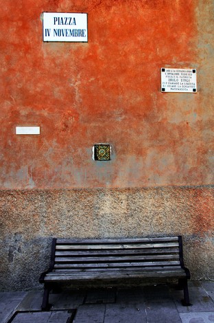 Lapide commemorativa su un muro arancione con panchina in piazza IV Novembre.
