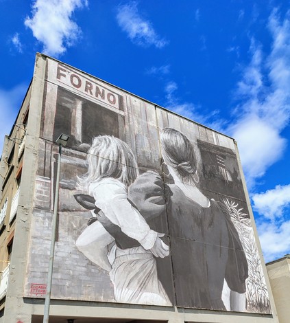 Murale che ritrae due figure femminili su un edificio, con cielo azzurro e nuvole bianche sullo sfondo.
