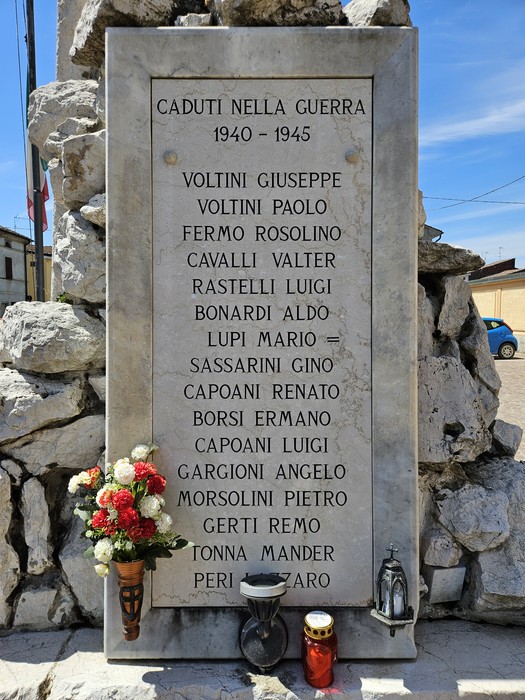 Monumento ai Caduti con nomi incisi, circondato da rocce e decorato con fiori e candele.
