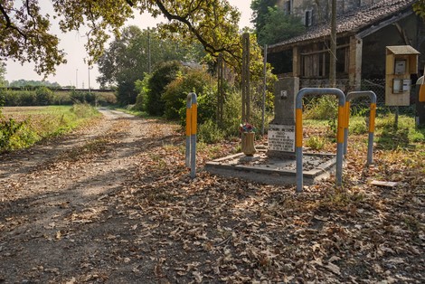 Cippo commemorativo immerso nella natura lungo una strada di campagna, circondato da alberi e foglie cadute.