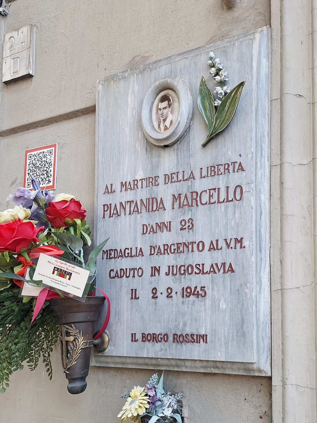 Lapide marmorea su parete, con ritratto fotografico, ramoscello e iscrizione dedicatoria, fiori deposti sotto.