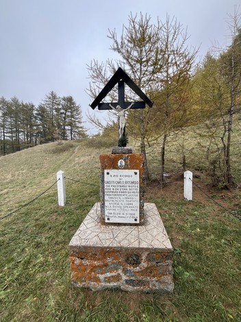 Monumento a Ghiotti, con una croce e una statua, circondato da un paesaggio naturale e alberi.