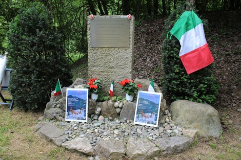 Cippo commemorativo con bandiera italiana, fiori e manifesti nel verde, dedicato agli eventi storici del luogo.