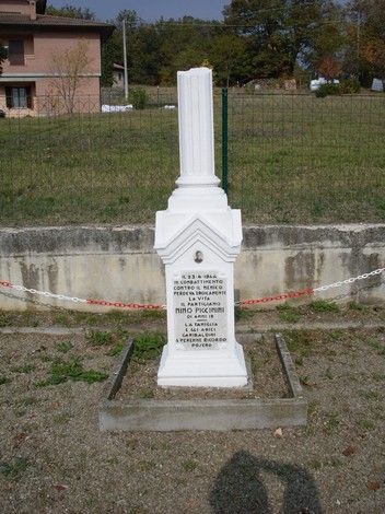 Cippo commemorativo dedicato a Nino Piccinini, in un'area rurale circondata da natura.