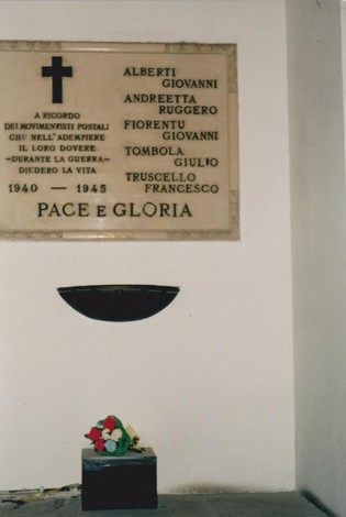 Lapide commemorativa in onore dei postali caduti, con una corona di fiori ai piedi e scritte in memoria.