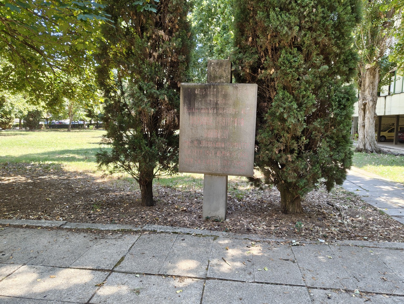 Stele in pietra con iscrizione, su basamento, tra alberi di cipresso in un'area verde.
