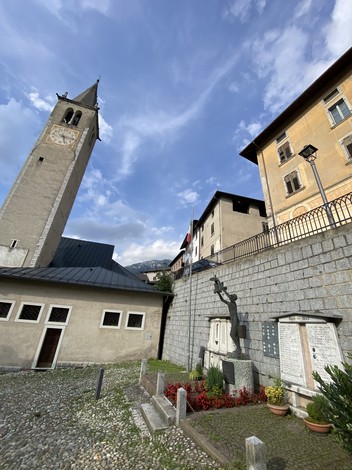 Monumento ai caduti di Castello Tesino, con torre campanaria e muratura in pietra sullo sfondo.