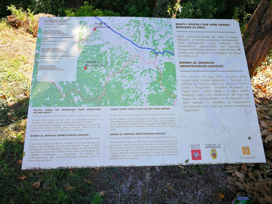 Pannello informativo con mappa e testi in più lingue, su supporto metallico immerso nel verde.
