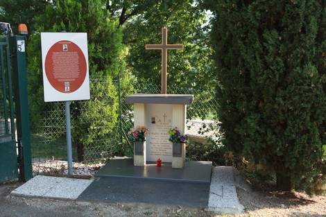 Cippo commemorativo con croce, fiori e pannello informativo, immerso nel verde.