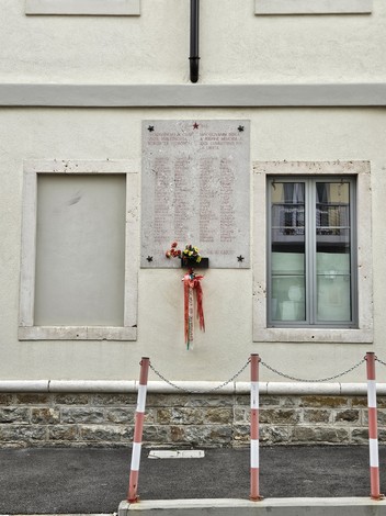 Lapide commemorativa in onore dei caduti per la libertà, con fiori e nastri colorati, su un muro bianco.