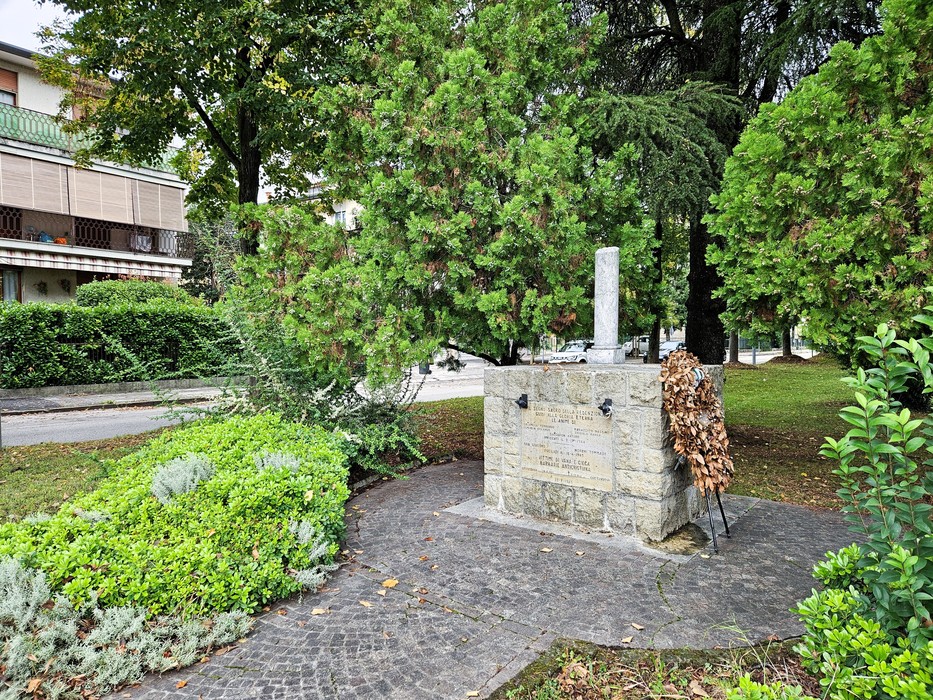 Cippo in pietra con iscrizione e colonnina, in un'aiuola tra alberi e siepi, con una corona secca appoggiata accanto.
