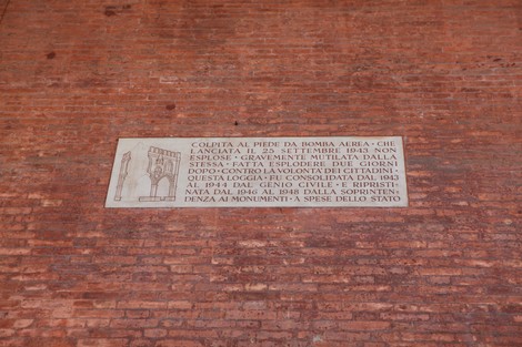 Lapide commemorativa che ricorda i danni causati da una bomba aerea nel 1943, situata su un muro di mattoni.