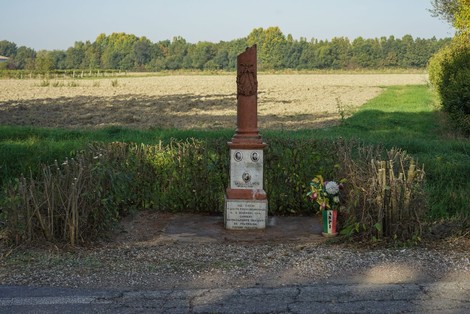 Cippo commemorativo circondato da vegetazione e fiori, situato in un'area rurale tranquilla.