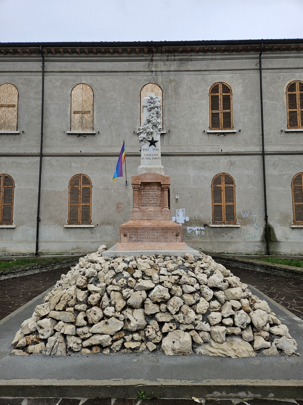 Monumento ai Caduti di Tabellano, con base in pietra e folto strato di sassi, sullo sfondo un edificio storico.
