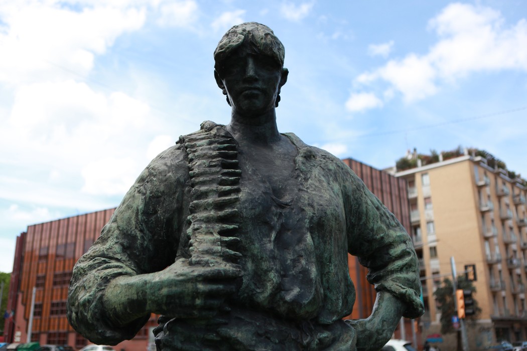 Statua di un partigiano con fucile, simbolo della Resistenza, sullo sfondo di edifici moderni e cielo blu.
