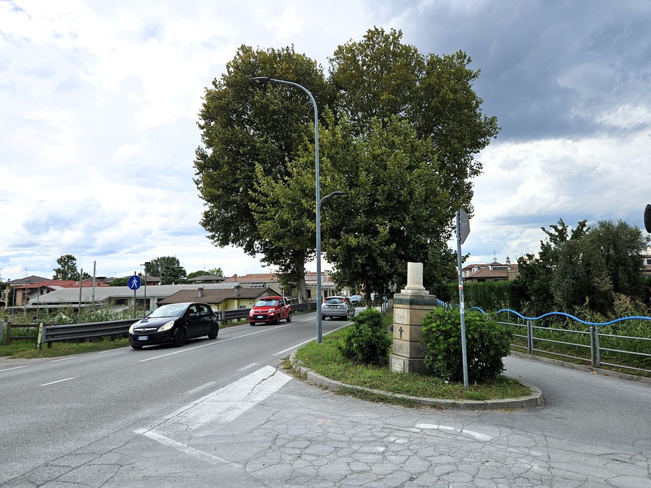 Cippo in pietra con iscrizione su un'aiuola spartitraffico, tra alberi e strade con auto in transito.