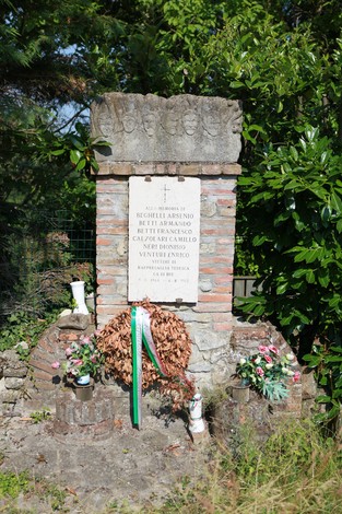 Monumento ai partigiani di Ca' di Bue, con una corona di fiori e una targa commemorativa. Verdeggianti alberi circostanti.