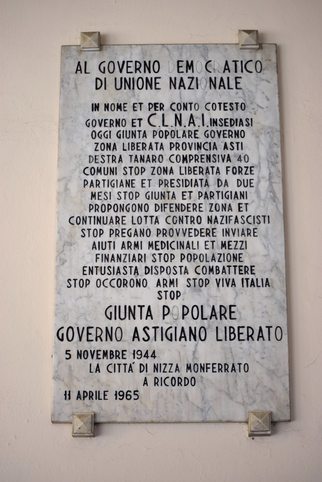 Lapide in marmo con lunga iscrizione, fissata a una parete con supporti angolari metallici.