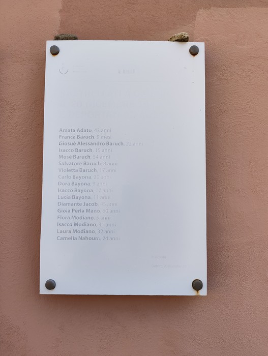 Targa bianca con elenco di nomi di deportati, fissata con quattro rosette metalliche a una parete.