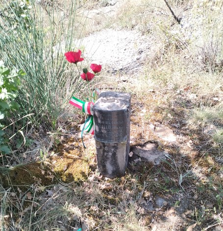 Cippo commemorativo a Misuri, con fiori e un nastro tricolore, immerso nella natura.