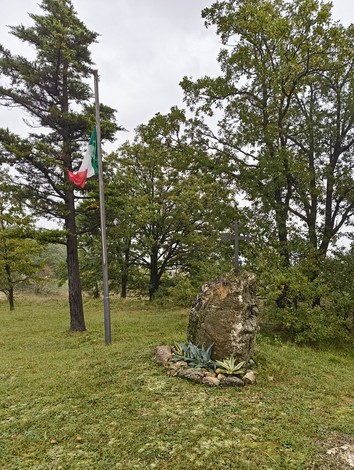 Monumento commemorativo per l'eccidio di Pian di Setta, circondato da vegetazione e bandiera italiana.