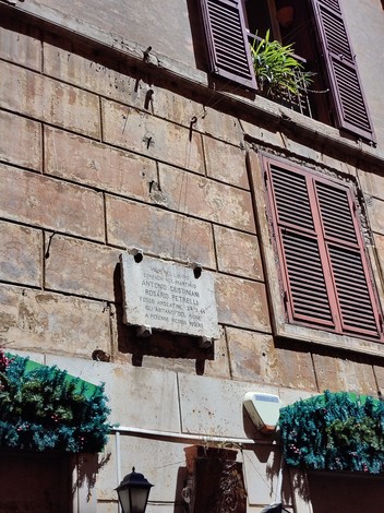 Lapide commemorativa su un muro, con decorazioni festive e finestre con persiane in legno.