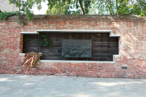 Monumento commemorativo con una scultura su una parete di mattoni, circondato da vegetazione e decorato con fiori.