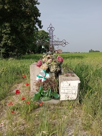 Cippo commemorativo di Scaramuzza, con fiori e decorazioni, immerso nel verde di un campo.
