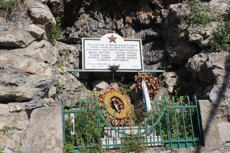 Lapide commemorativa ai caduti per la Libertà, incastonata tra le rocce, con una corona d'alloro e decorazioni floreali.