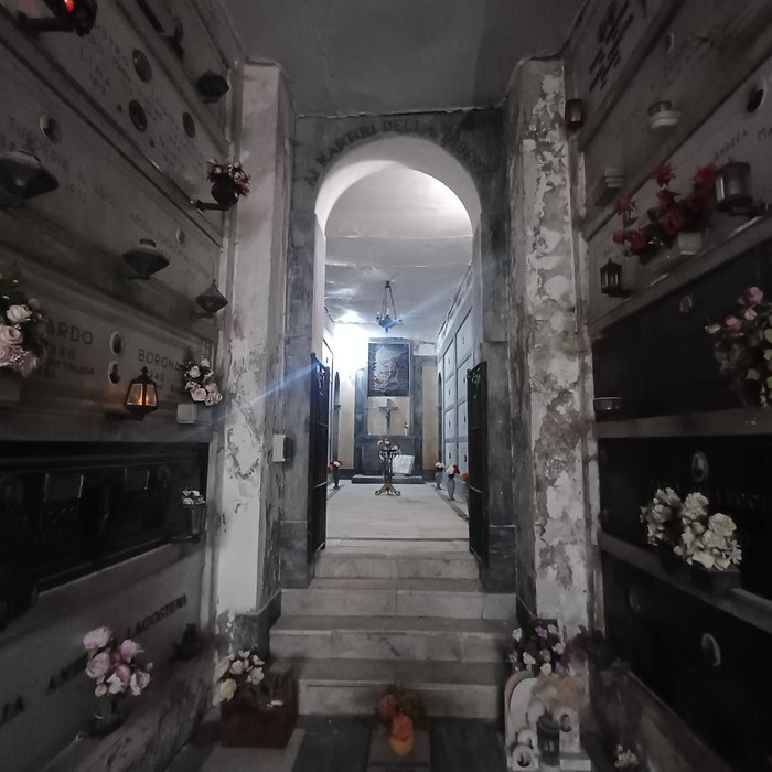 Interno di un sacrario con loculi marmorei lungo le pareti, corridoio verso un altare con fiori e lumini.