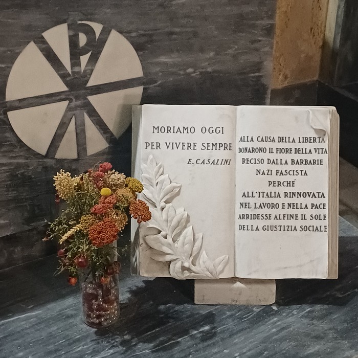 Libro aperto in marmo con iscrizione e decorazione a foglie, su leggio, accanto a un vaso di fiori secchi.