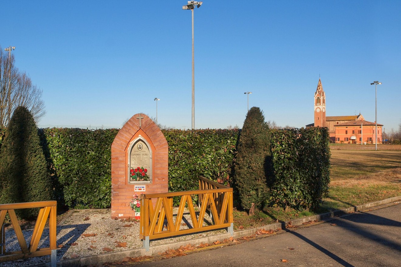 Monumento a Gilioli circondato da siepi, con chiesa in lontananza e cielo sereno.