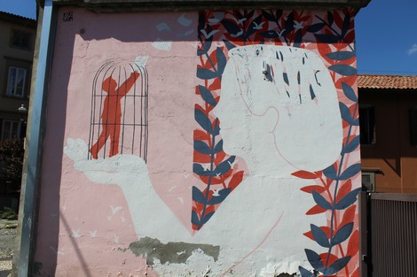 Murale colorato su un muro, raffigurante una figura che tiene una gabbia con un bambino e foglie decorative.