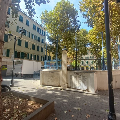 Monumento in pietra con lapidi incise, recintato, in una piazza alberata circondata da edifici.