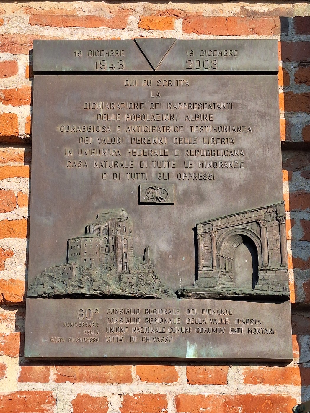 Targa in bronzo su parete di mattoni, con iscrizione e rilievi di un castello e un arco trionfale.