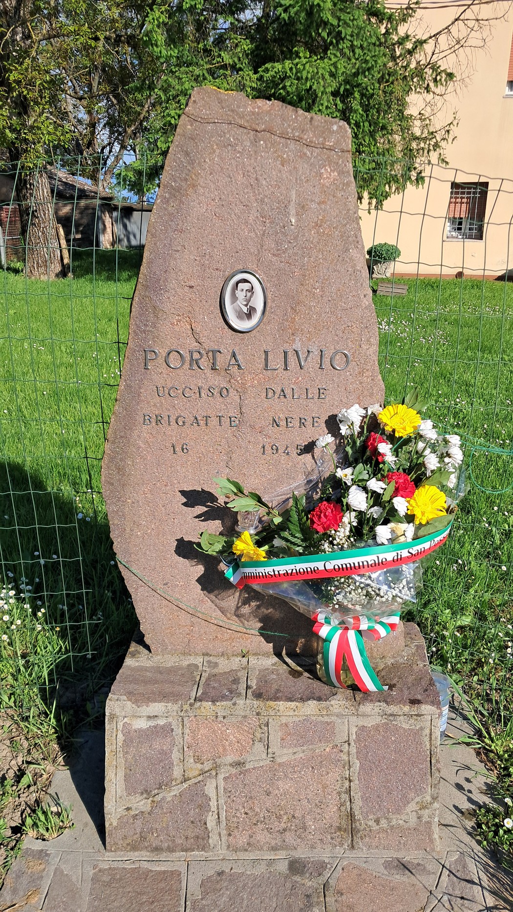 Monumento in pietra dedicato a Porta Livio, con fiori colorati e un nastro tricolore.