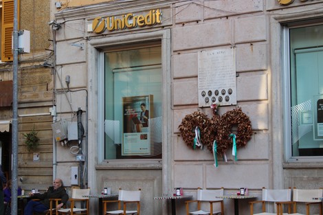 Lapide commemorativa con decorazioni floreali, affissa su un muro accanto a una banca, in un ambiente urbano.