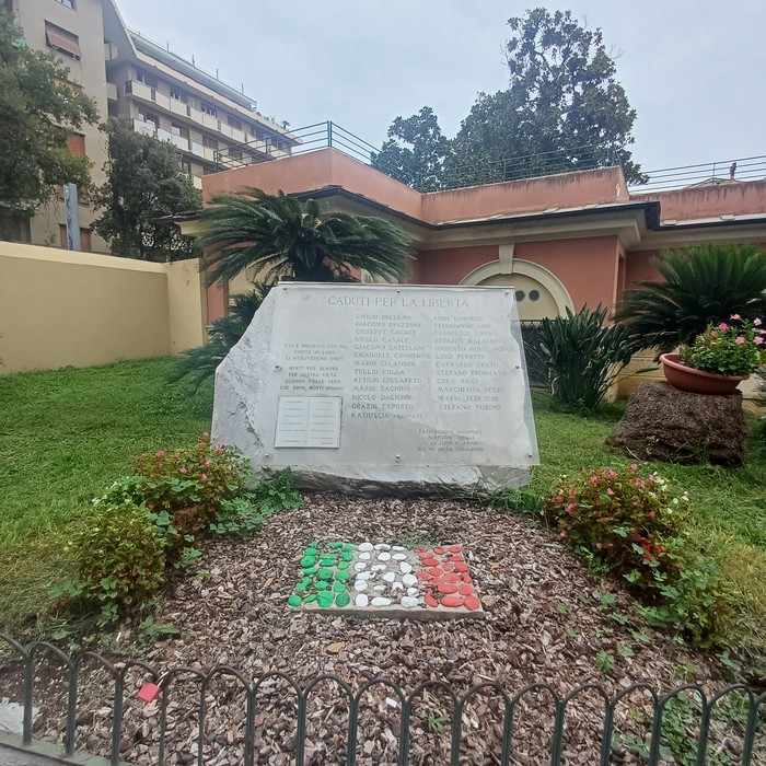 Lapide in marmo con iscrizione, in un giardino, davanti a un mosaico con i colori della bandiera italiana.
