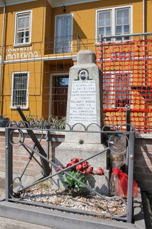 Cippo commemorativo a Malavasi, circondato da fiori, con una struttura architettonica colorata sullo sfondo.