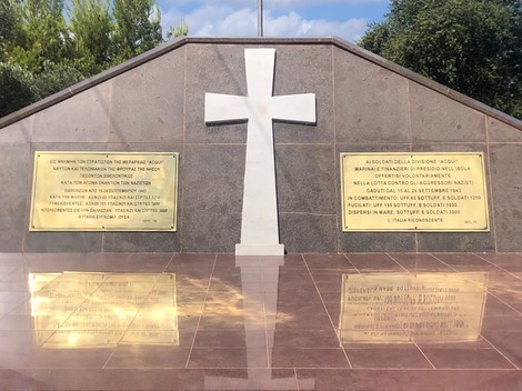Monumento del "Sacrario dei caduti di Cefalonia", con croce bianca e placca commemorativa, circondato da vegetazione.