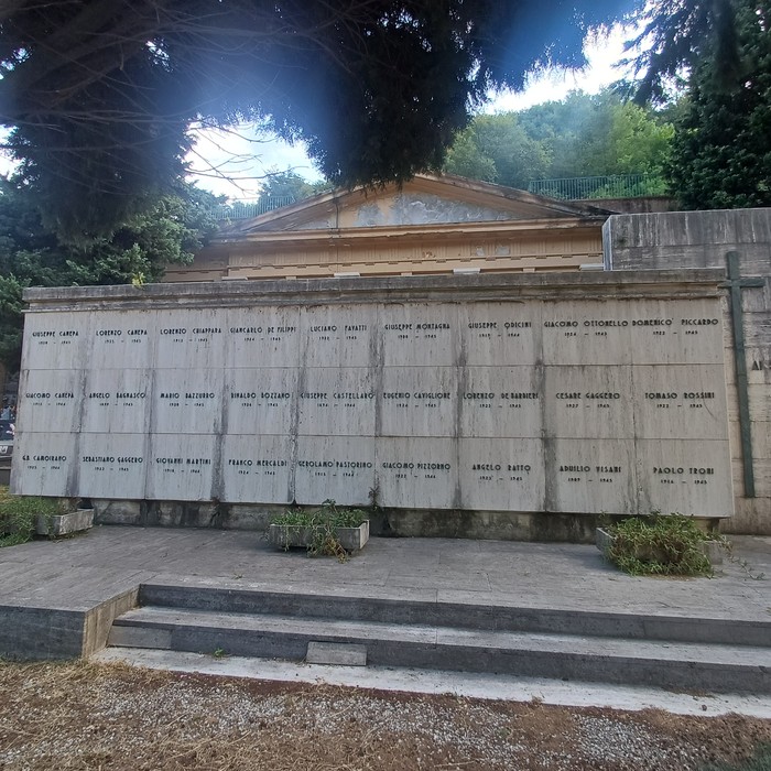 Parete con loculi marmorei e iscrizioni, sormontata da un frontone triangolare, tra gli alberi.