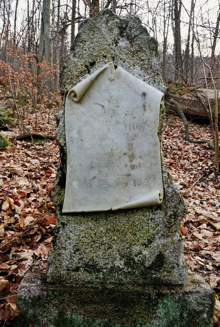 Cippo in pietra nel bosco, con lapide ovale incisa, parzialmente consumata, tra foglie secche.