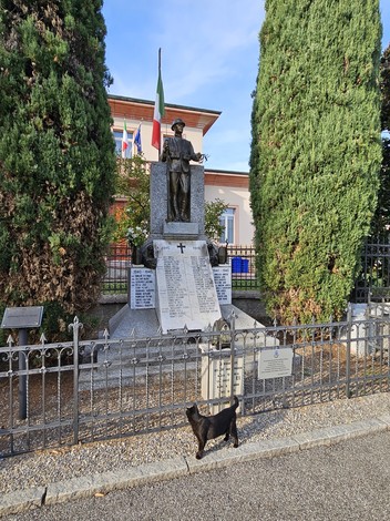 Monumento in pietra con statua bronzea su piedistallo con iscrizione, tra due cipressi, recinzione in ferro.