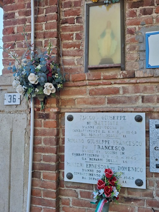 Lapide in marmo con iscrizione su muro di mattoni, sotto una nicchia con statua della Madonna e fiori.