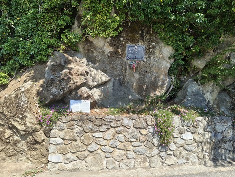 Targa con iscrizione su parete rocciosa, fiori deposti sotto, muretto in pietra e cartello informativo nei pressi.