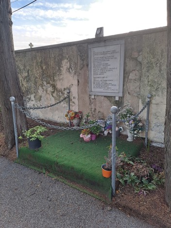 Lapide commemorativa con fiori e decorazioni, situata accanto a un muro, tra cui piante adornano l'area.