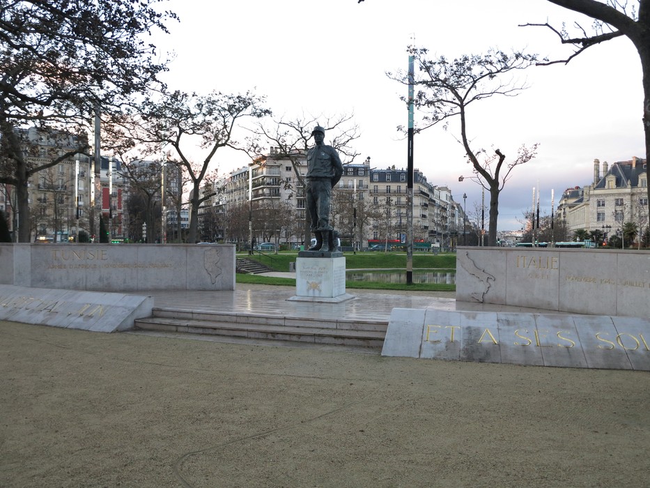 Statua di Juin in un parco urbano, circondata da alberi e edifici storici, con un'atmosfera di memoria e rispetto.