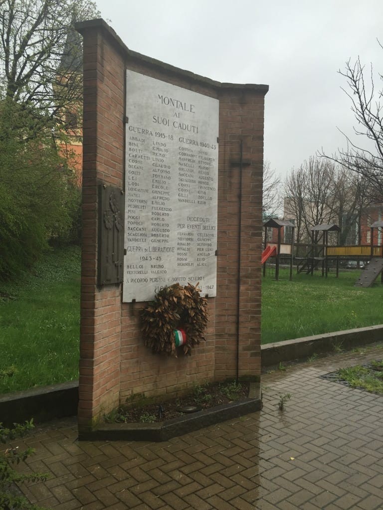 Monumento in mattoni con lapide di marmo e iscrizione, corona fiorita alla base, in un'area verde.